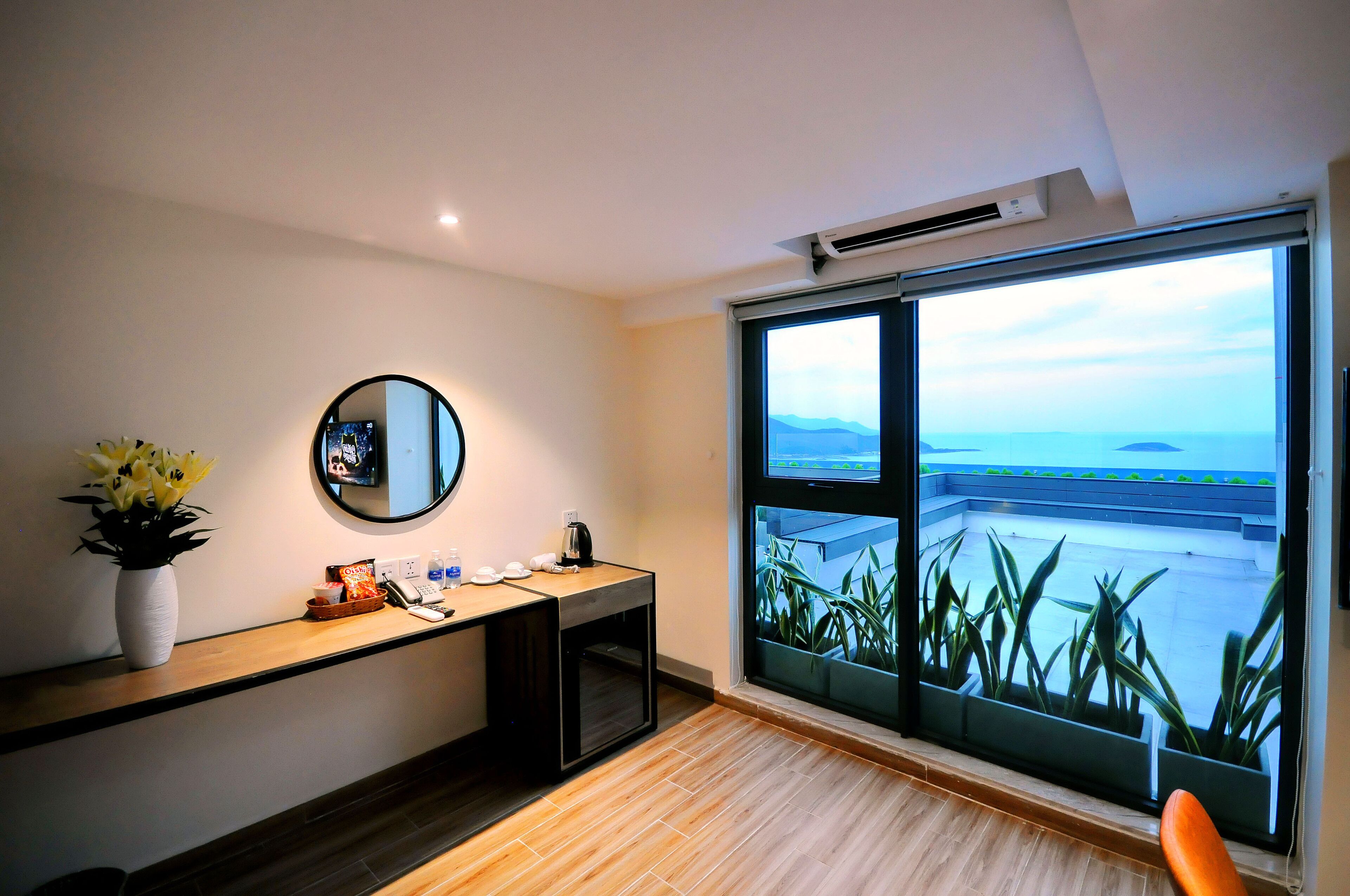 A hotel room with a wooden desk, a mirror, a vase with flowers, a coffee maker, and a large window with a view of a balcony and greenery.