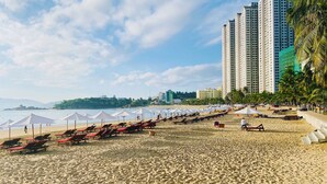 Ein Strand mit Liegestühlen und Sonnenschirmen, eine von Palmen gesäumte Uferpromenade und hohe Gebäude im Hintergrund.