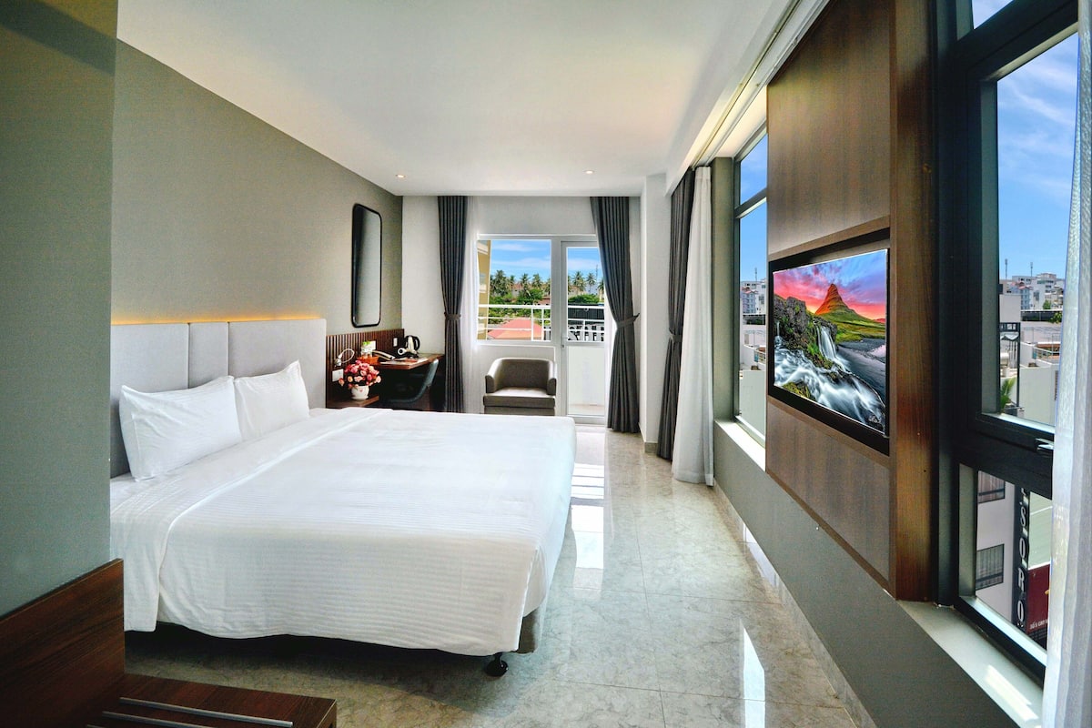 Une chambre d’hôtel avec un grand lit, un bureau avec une chaise, une vue sur le paysage urbain à travers de grandes fenêtres et un balcon avec une balustrade.