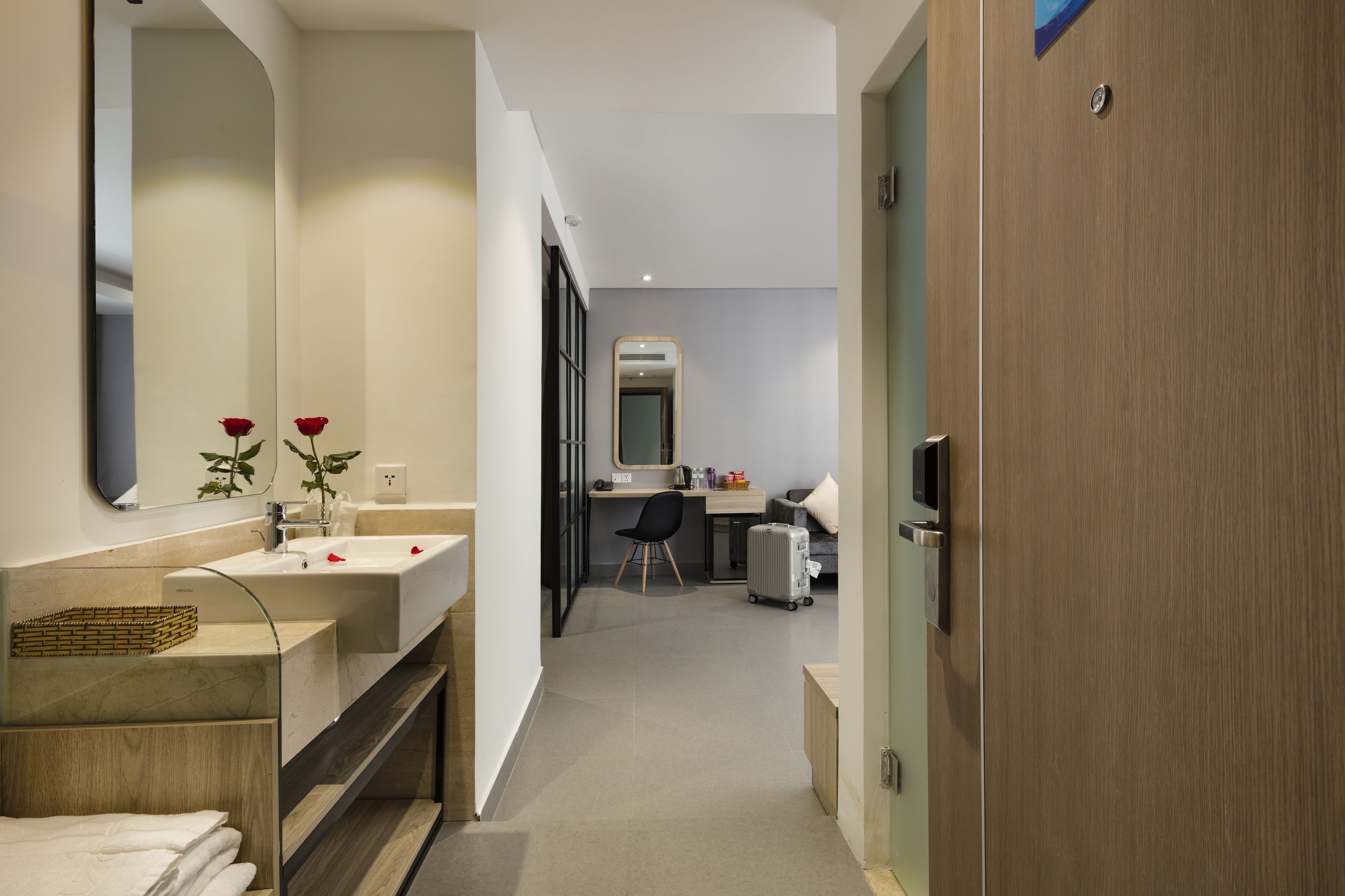 A modern bathroom with a large mirror, a sink, and a vanity. There is a wooden door on the right and a desk with a chair in the background.