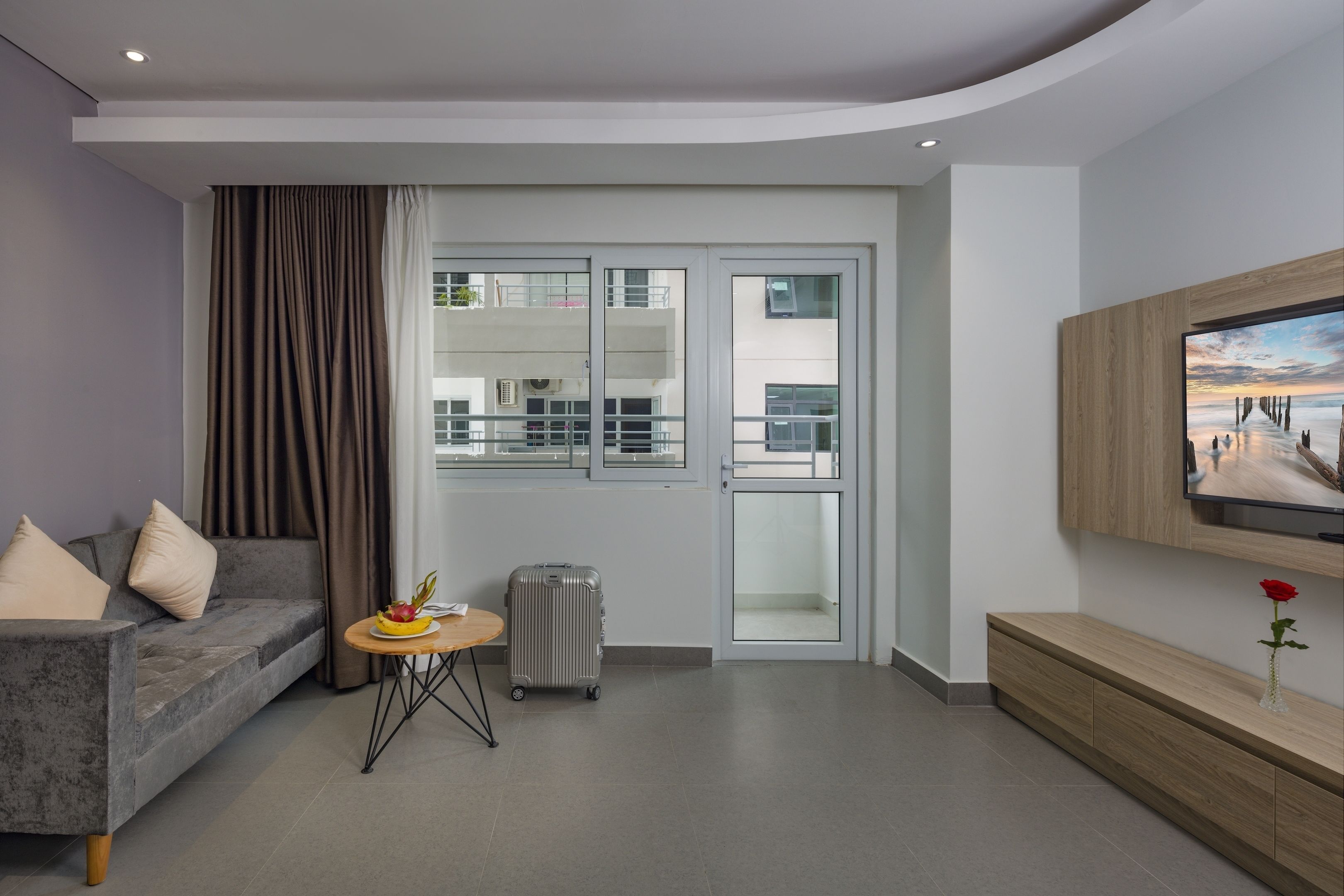 Une chambre d’hôtel moderne avec un canapé gris, une petite table ronde sur laquelle se trouve un panier de fruits, une valise, une console en bois ornée d’un vase contenant une rose rouge, et un téléviseur fixé au mur.