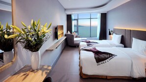Ein modernes Hotelzimmer mit großem Fenster und Meerblick, zwei Betten, einer Sitzecke und einem Blumenstrauß.
