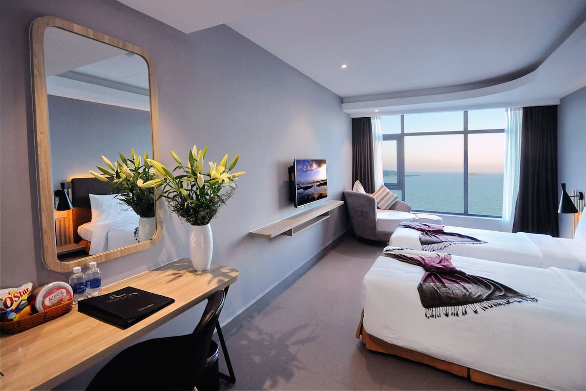 Une chambre d’hôtel avec un grand miroir, un bureau avec un panier de collations, une plante, une télévision, deux lits, une fenêtre donnant sur la mer et une petite étagère.