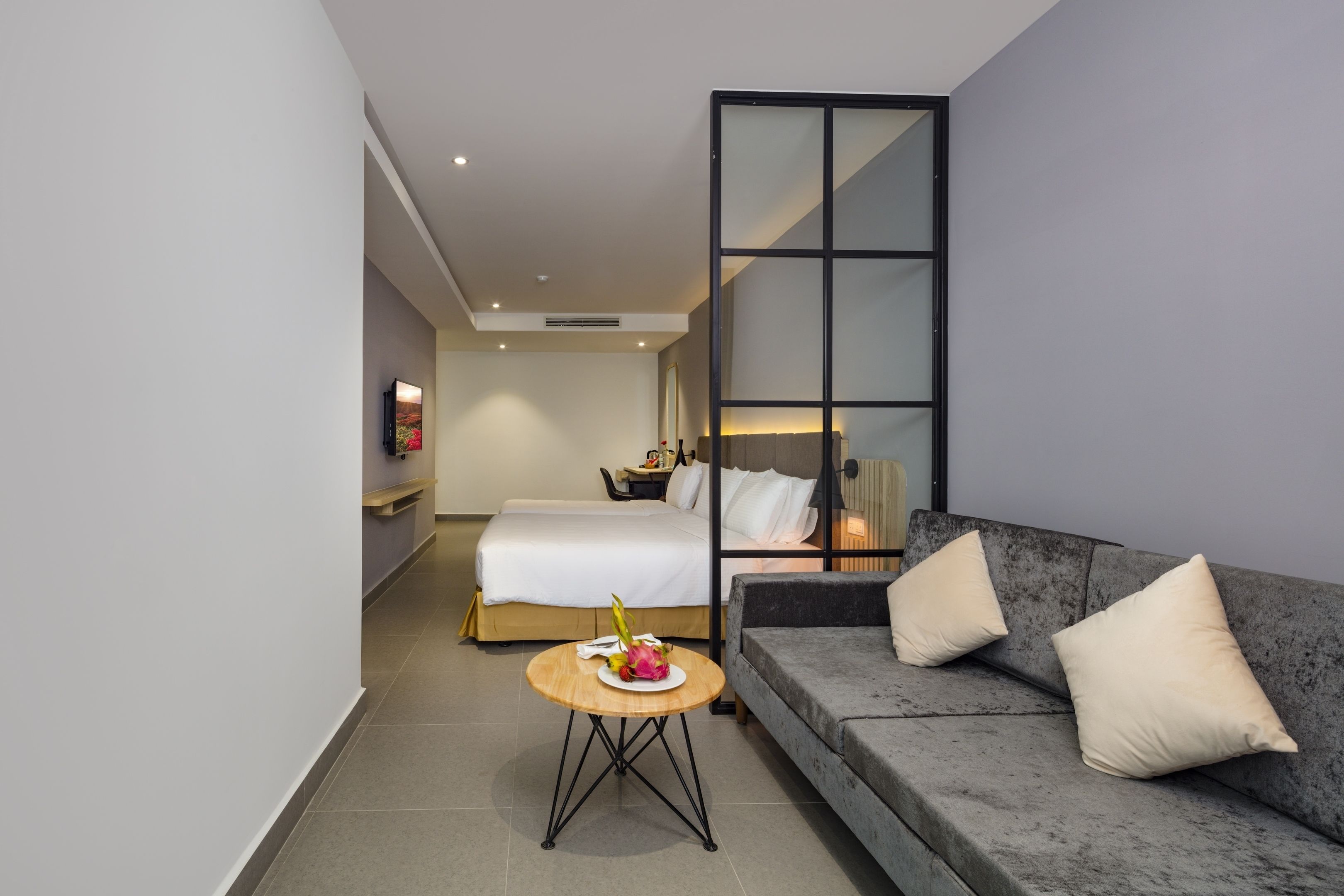 Une chambre d’hôtel moderne, équipée d’un lit, d’un canapé, d’une petite table ronde sur laquelle se trouve une assiette de fruits, et d’une grande fenêtre offrant une vue sur la ville.