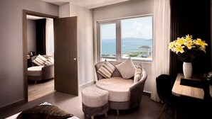 Ein modernes Wohnzimmer mit Sofa, Hocker und Esstisch. Ein großes Fenster bietet Blick auf das Meer und die Berge.