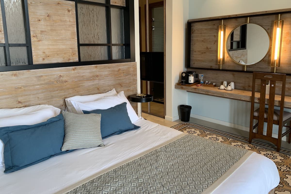 Une chambre d’hôtel avec un grand lit, une tête de lit en bois, une table de chevet avec un miroir et un petit bureau équipé d’une machine à café.