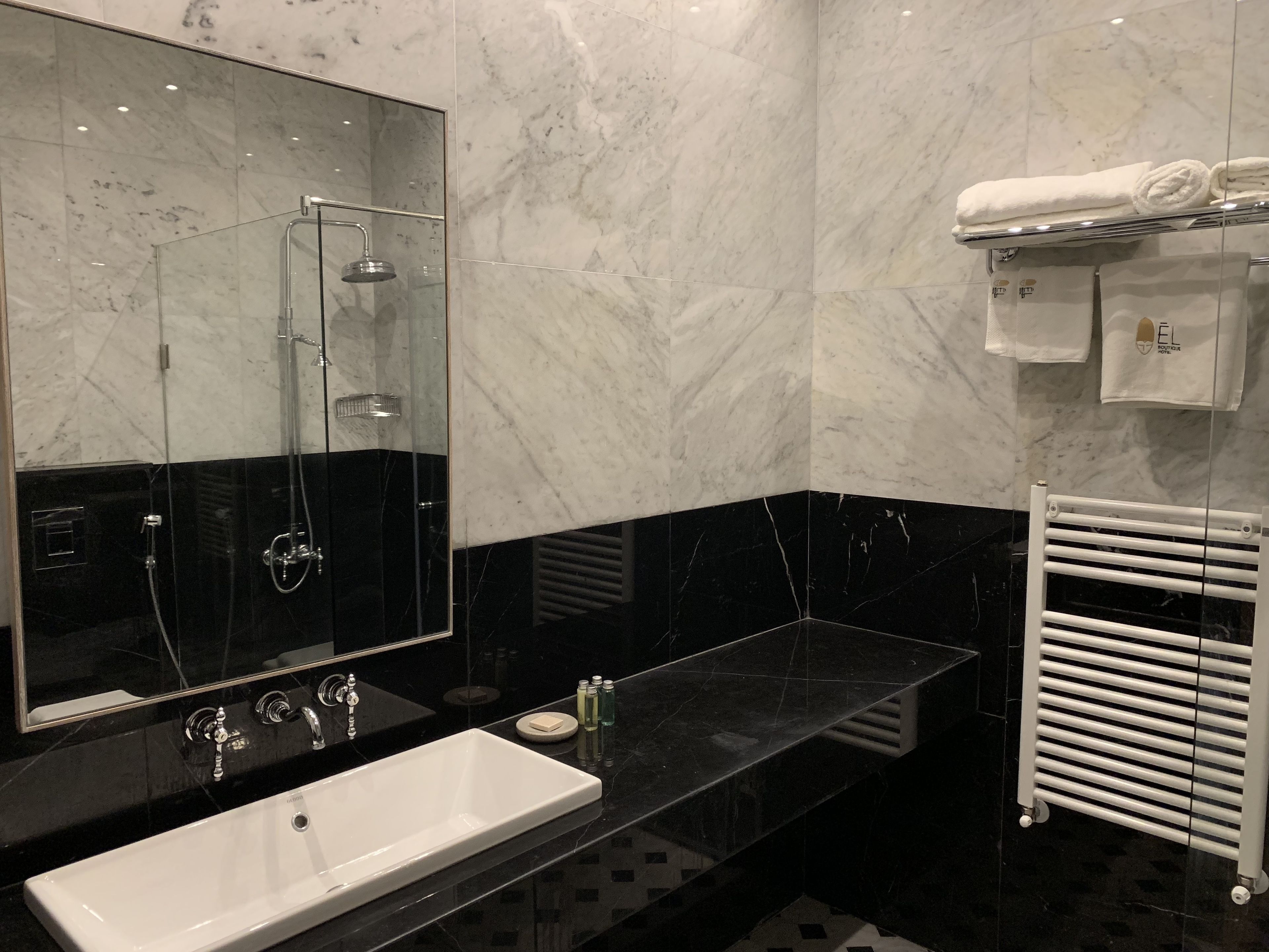 A modern bathroom with a large mirror, a black sink, a shower, and a towel rack with towels.