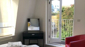 Ein Schlafzimmer mit Bett, Schreibtisch, einem roten Sessel und einem Balkon mit Blick auf Grünflächen.