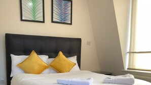 Ein ordentlich bezogenes Bett mit weißen Bettwäsche und zwei gelben Kissen, ein schwarzes Kopfstück und zwei gerahmte abstrakte Gemälde an der Wand.