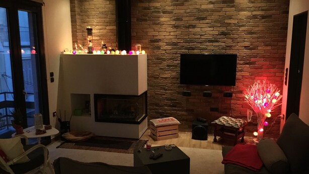 A modern living room with a fireplace, a flat-screen TV mounted on a brick wall, and a cozy seating area. - Bright Cozy vintage apartment (Athina)