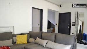 A modern living room with a grey sectional sofa, a wooden coffee table, and a yellow throw pillow. There are two doors, one leading to a staircase and another to a different room. A ladder is leaning against the wall, and a sign indicates the room is checked by SPOTAHOME. - Bright Cozy vintage apartment (Athina)
