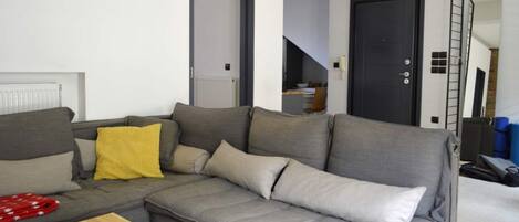 A modern living room with a grey sectional sofa, a wooden coffee table, and a yellow throw pillow. There are two doors, one leading to a staircase and another to a different room. A ladder is leaning against the wall, and a sign indicates the room is checked by SPOTAHOME.