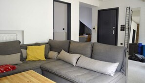 A modern living room with a grey sectional sofa, a wooden coffee table, and a yellow throw pillow. There are two doors, one leading to a staircase and another to a different room. A ladder is leaning against the wall, and a sign indicates the room is checked by SPOTAHOME.