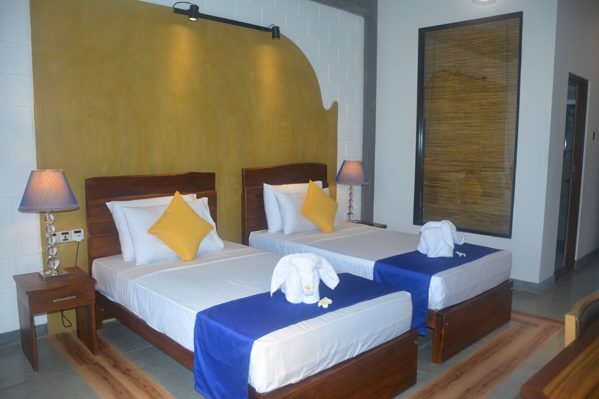 A hotel room with two beds, each with a wooden headboard, white bedding, and a blue accent. There are lamps on nightstands, a large mirror, and a wooden floor.