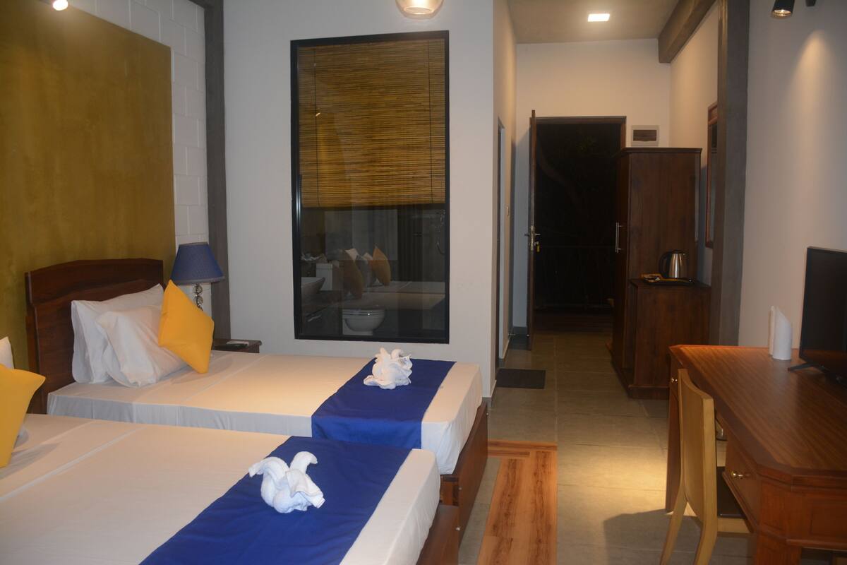 A hotel room with two beds, a wooden desk, a television, and a large window with blinds.