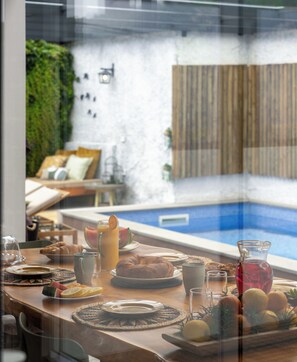 A dining table with breakfast items, a pool, and a view of a garden and outdoor seating area.