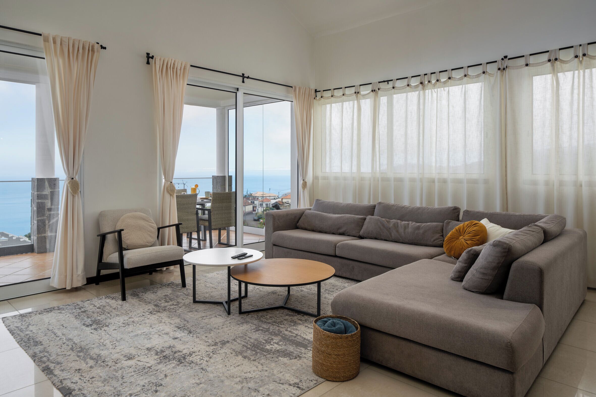 A modern living room with a grey sofa, a round coffee table, and a view of the sea through large windows.