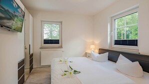 Une chambre à coucher avec un lit, deux fenêtres, un téléviseur, un radiateur et une table de chevet avec une lampe.