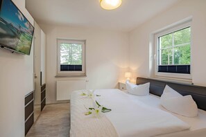 Une chambre à coucher avec un lit, deux fenêtres, un téléviseur, un radiateur et une table de chevet avec une lampe.