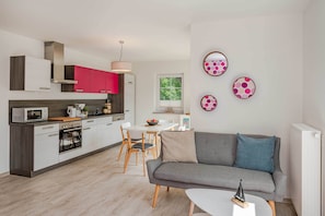 A modern kitchen with white and red cabinets, a dining area with a white table and chairs, and a grey sofa with a blue pillow.