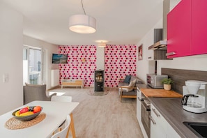 A modern kitchen with a coffee maker, a toaster, and a stove. There is a dining area with a table and chairs, and a living area with a sofa and a TV.