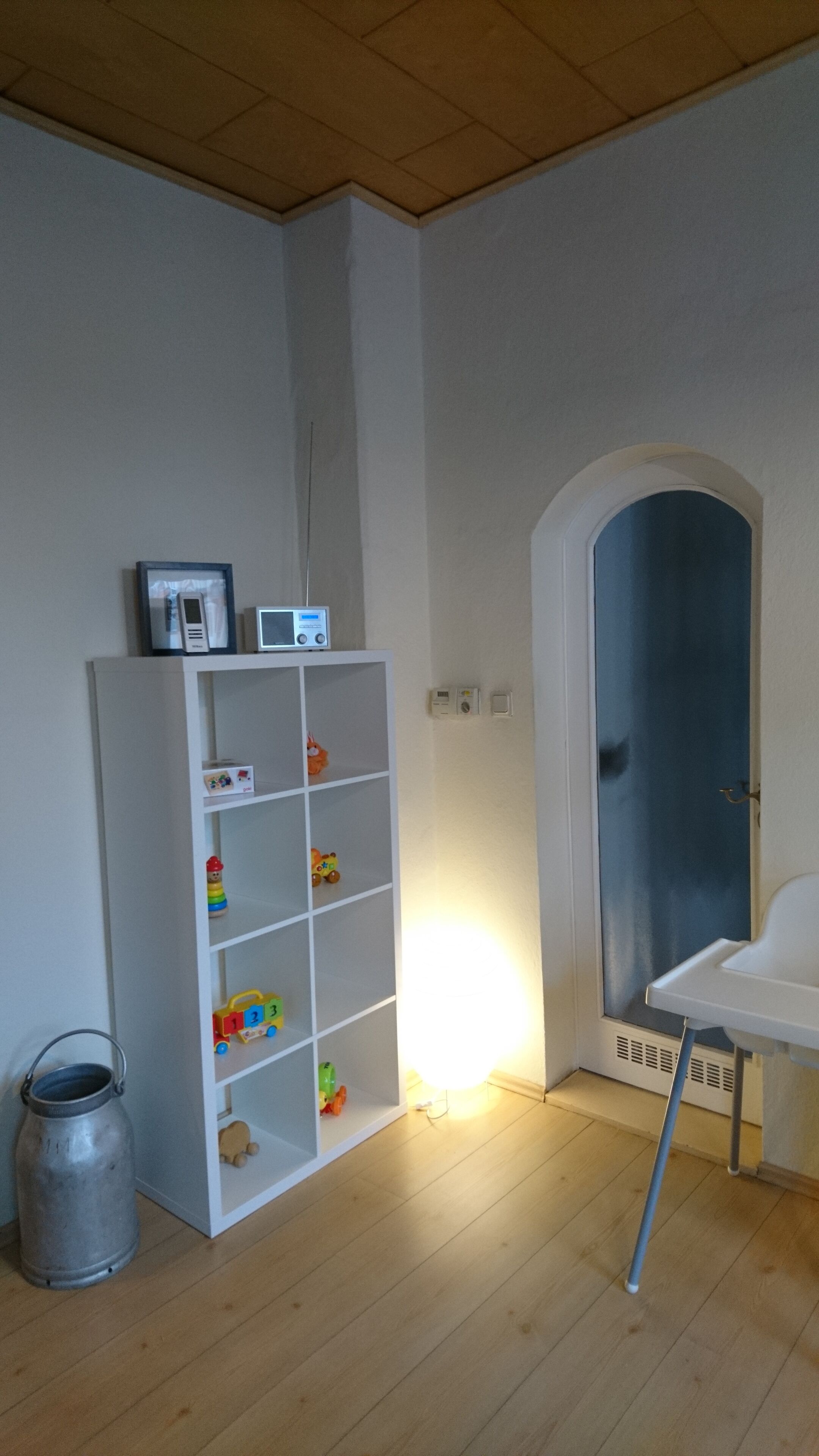 Une pièce avec une bibliothèque blanche, une petite table, un seau en métal et une porte surmontée d’un rideau bleu.
