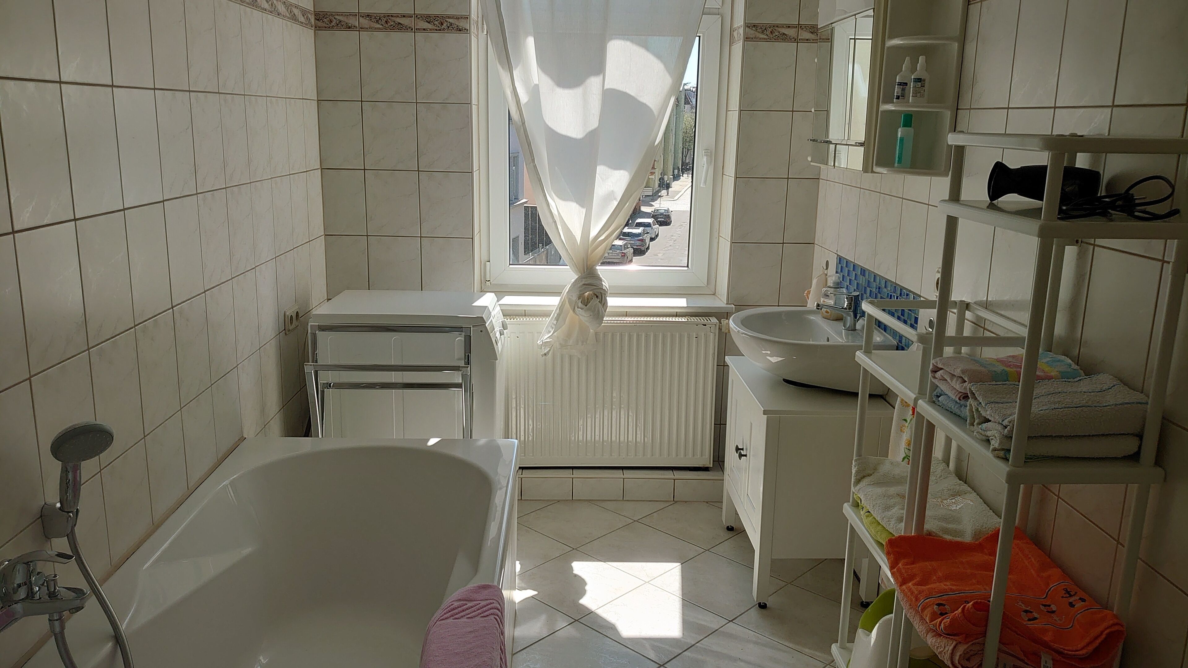 A bathroom with a bathtub, a window with a curtain, a sink, and a shelf with toiletries.