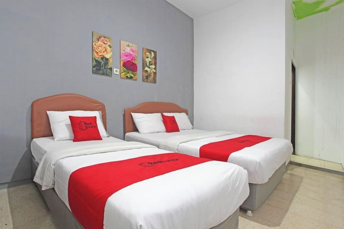 Een hotelkamer met twee bedden, elk met een rood-wit dekbed, en een grijze hoofdbord. Boven de bedden hangen drie bloemrijke schilderijen.