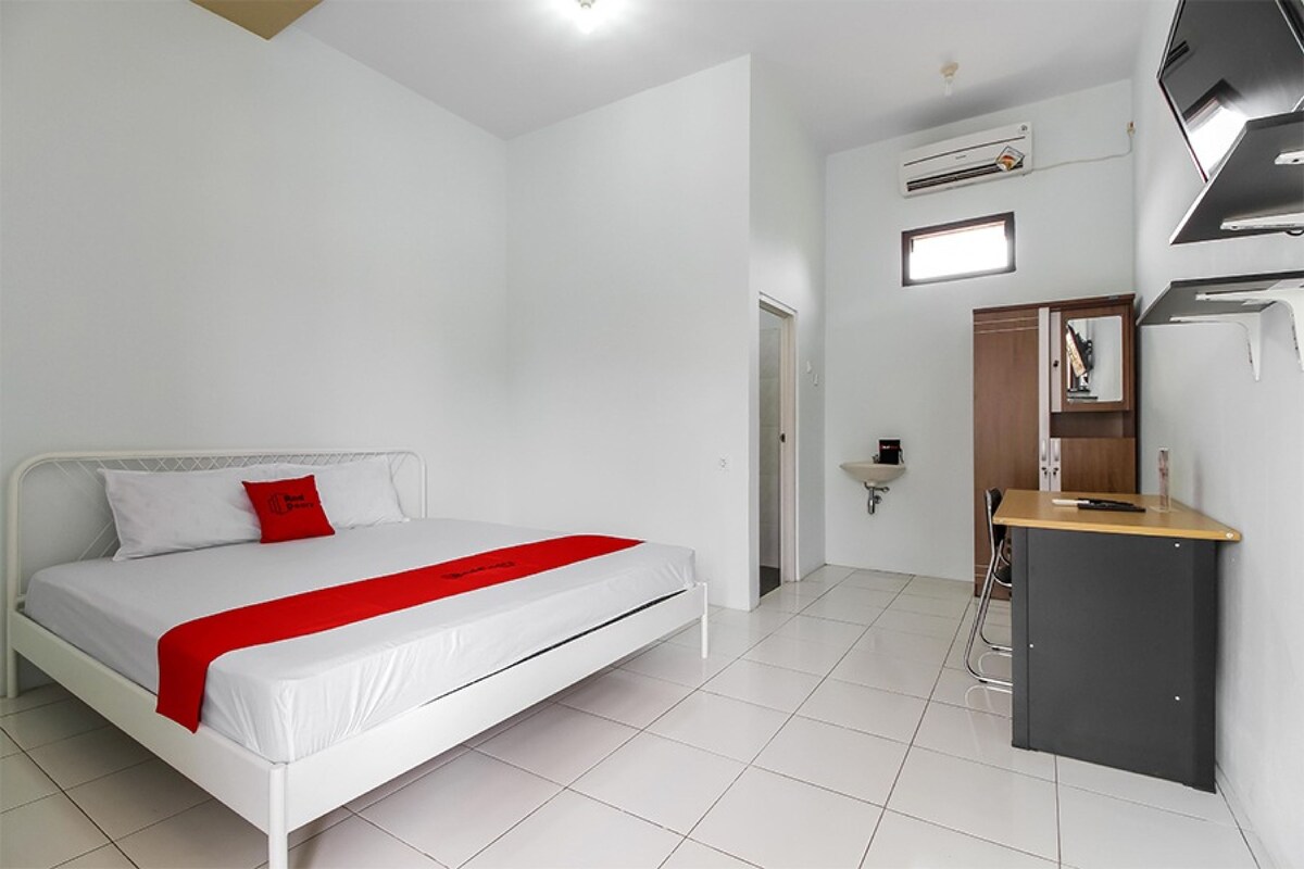 Una habitación de hotel moderna con cama blanca, almohada decorativa roja, escritorio pequeño y armario empotrado.