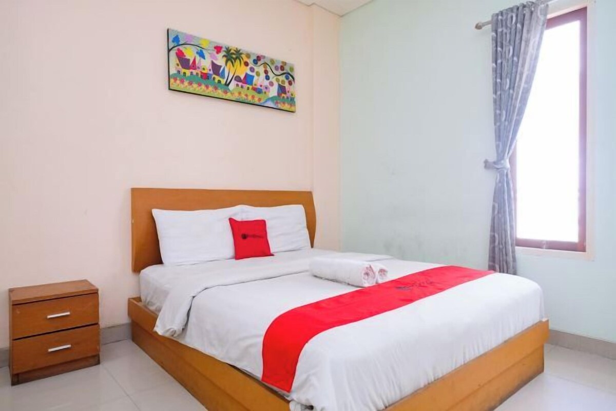 Una cama bien hecha con ropa blanca y un cojín rojo de acento, una mesita de noche de madera con cajones, un cuadro abstracto y colorido en la pared y una ventana con cortina.