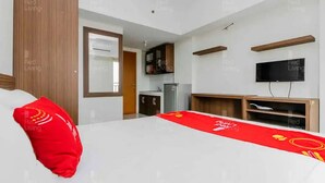 A hotel room with a bed, a red pillow, a TV mounted on the wall, and a small kitchenette. - RedLiving Apartemen Margonda Residence - Tower 2 (Depok)