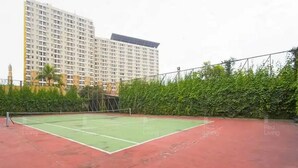 A tennis court with a net, surrounded by a red surface, and a tall building in the background. - RedLiving Apartemen Margonda Residence - Tower 2 (Depok)