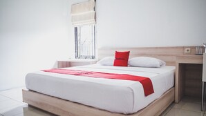 A modern hotel room with a wooden bed, white bedding, a red pillow, a window with a beige blind, and a small bedside table with a lamp. - RedDoorz near Alun-alun Kota Sukabumi (Sukabumi)