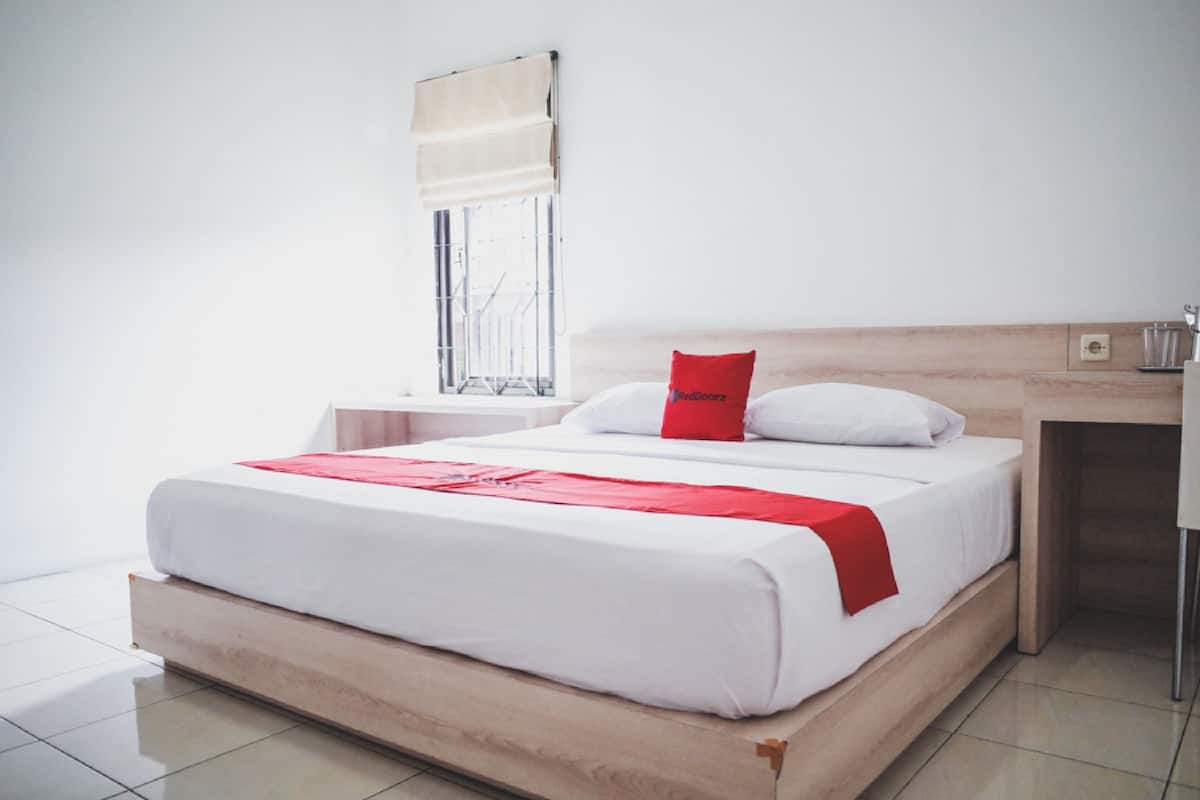 A modern hotel room with a wooden bed, white bedding, a red pillow, a window with a beige blind, and a small bedside table with a lamp.