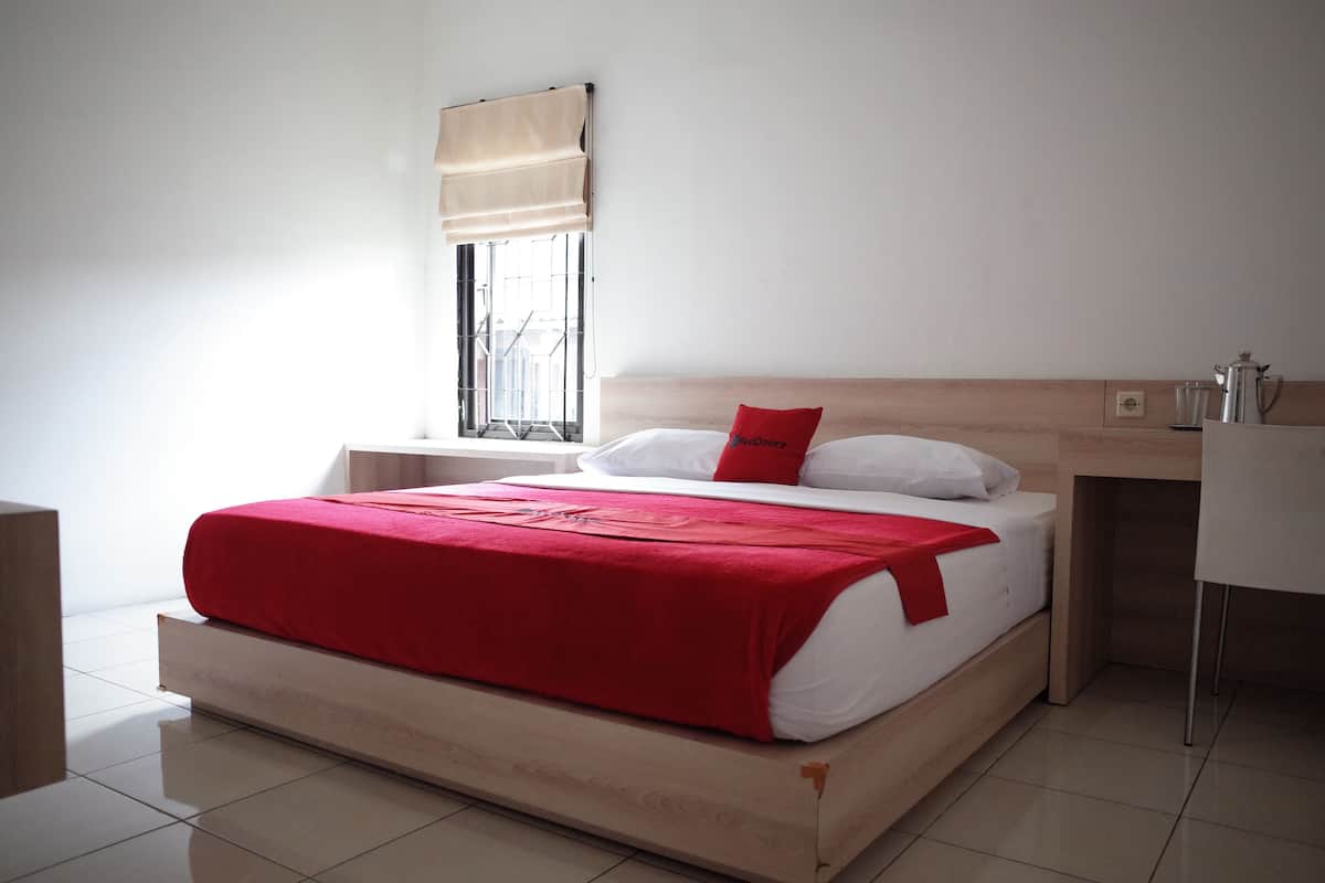 A modern hotel room with a wooden bed, a red blanket, a white pillow, a window with a beige blind, and a small desk with a cup and a kettle.