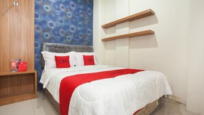 A neatly made bed with a white and red bedspread, two red pillows with a logo, a wooden nightstand with a red item, and a blue patterned wall.