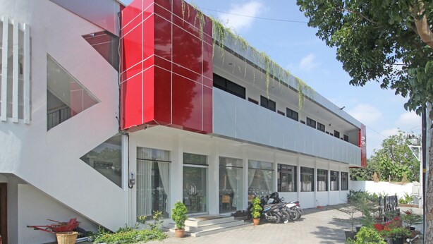 Un bâtiment moderne à la façade rouge et blanche, doté de grandes fenêtres et d’un espace d’entrée pavé agrémenté de plantes en pot et d’une moto.