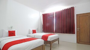 Une chambre d’hôtel avec deux lits, chacun agrémenté de draps blancs et de touches de couleur rouge, une porte en bois et une fenêtre avec des rideaux rouges.