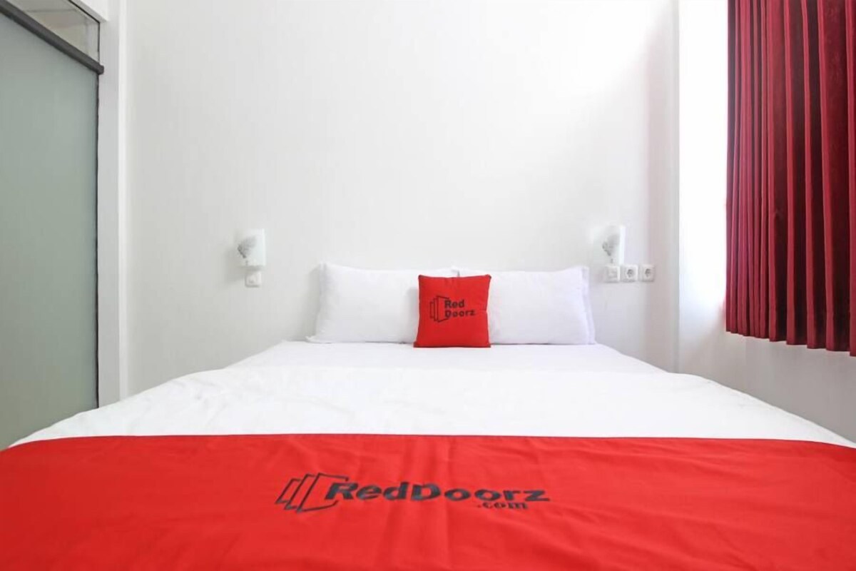Una habitación de hotel con una cama, un almohadón rojo con el logotipo 'RedDoorz', paredes blancas y una cortina roja.