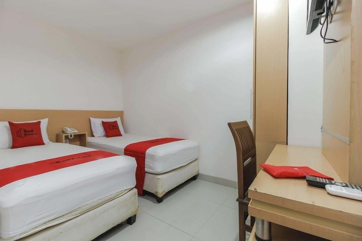 Habitación de hotel con dos camas, cada una con ropa de cama roja y blanca, un escritorio de madera y una silla. Hay un televisor montado en la pared y un teléfono en el escritorio.