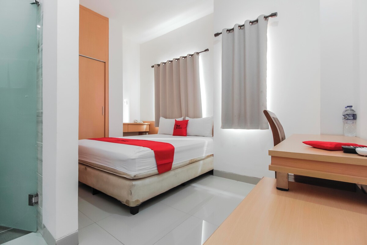 Habitación de hotel con cama, escritorio, silla, ventana con cortinas y puerta.
