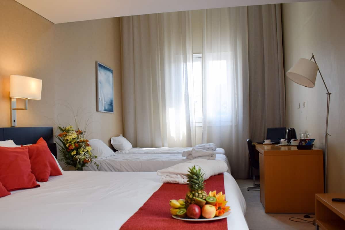 A hotel room with two beds, a desk, a lamp, a painting, and a fruit tray.