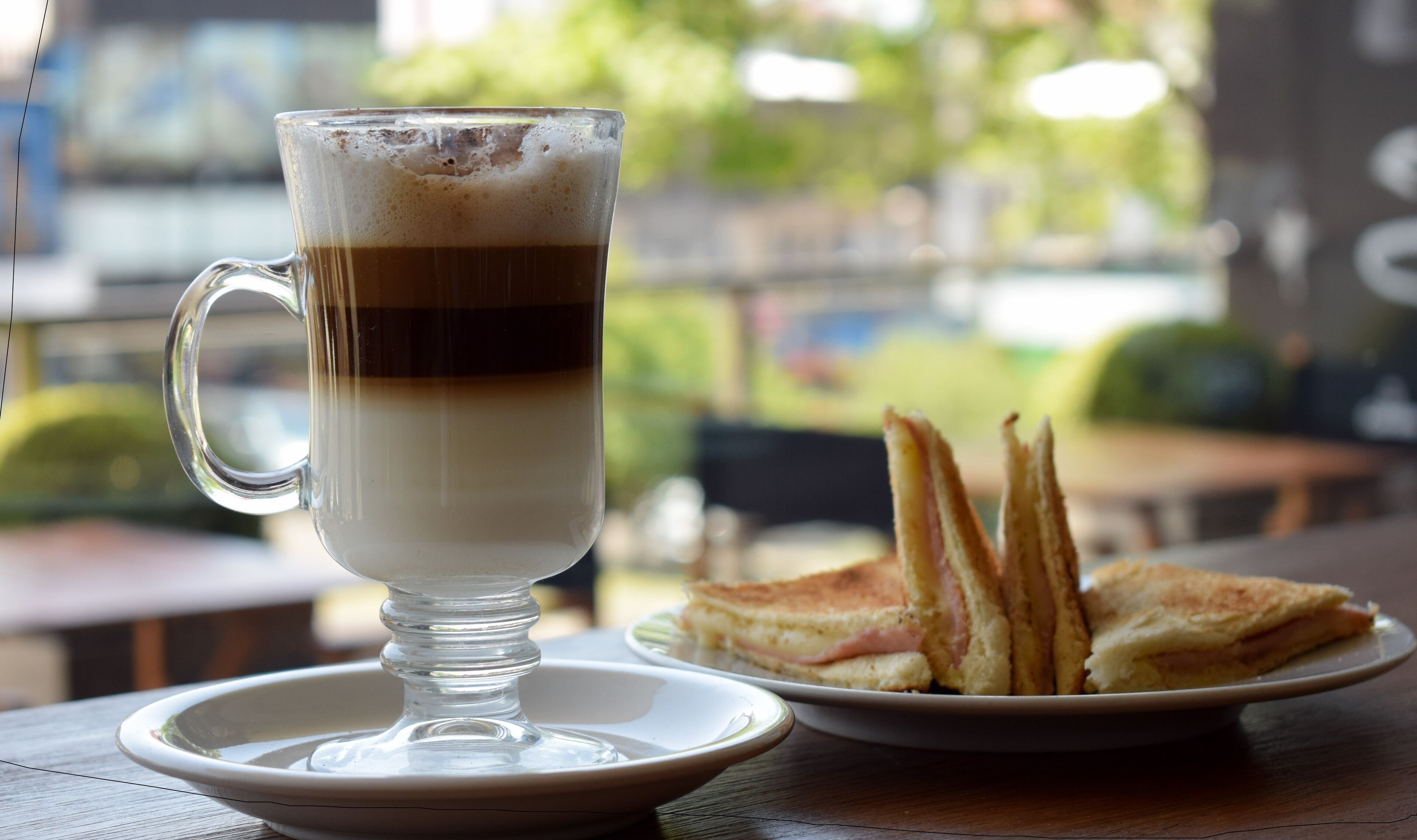 A clear glass cup with a layered beverage, a plate with sandwiches, and a wooden table.