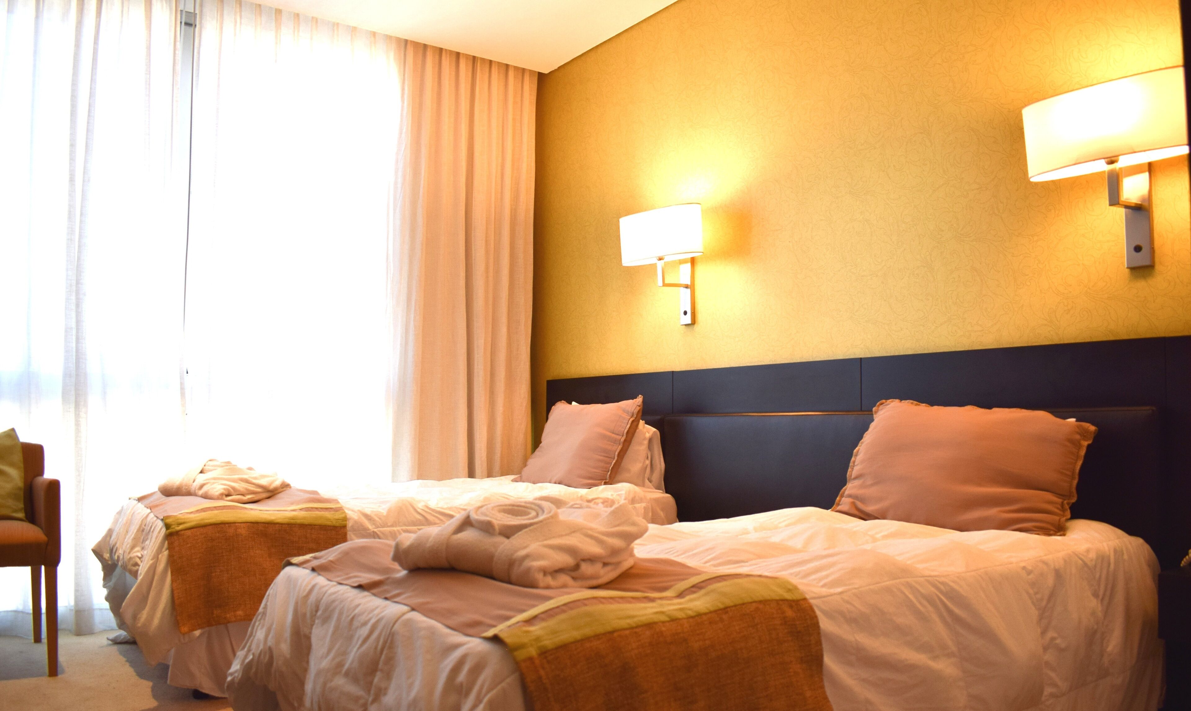 Habitación de hotel con dos camas, cabeceros, mesitas de noche, una lámpara y un ventanal con cortinas.