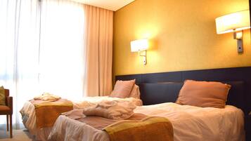 Habitación de hotel con dos camas, cabeceros, mesitas de noche, una lámpara y un ventanal con cortinas.