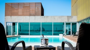 Un área de piscina con sillones de descanso, un cubo metálico con una botella, y un edificio moderno con amplios ventanales al fondo.