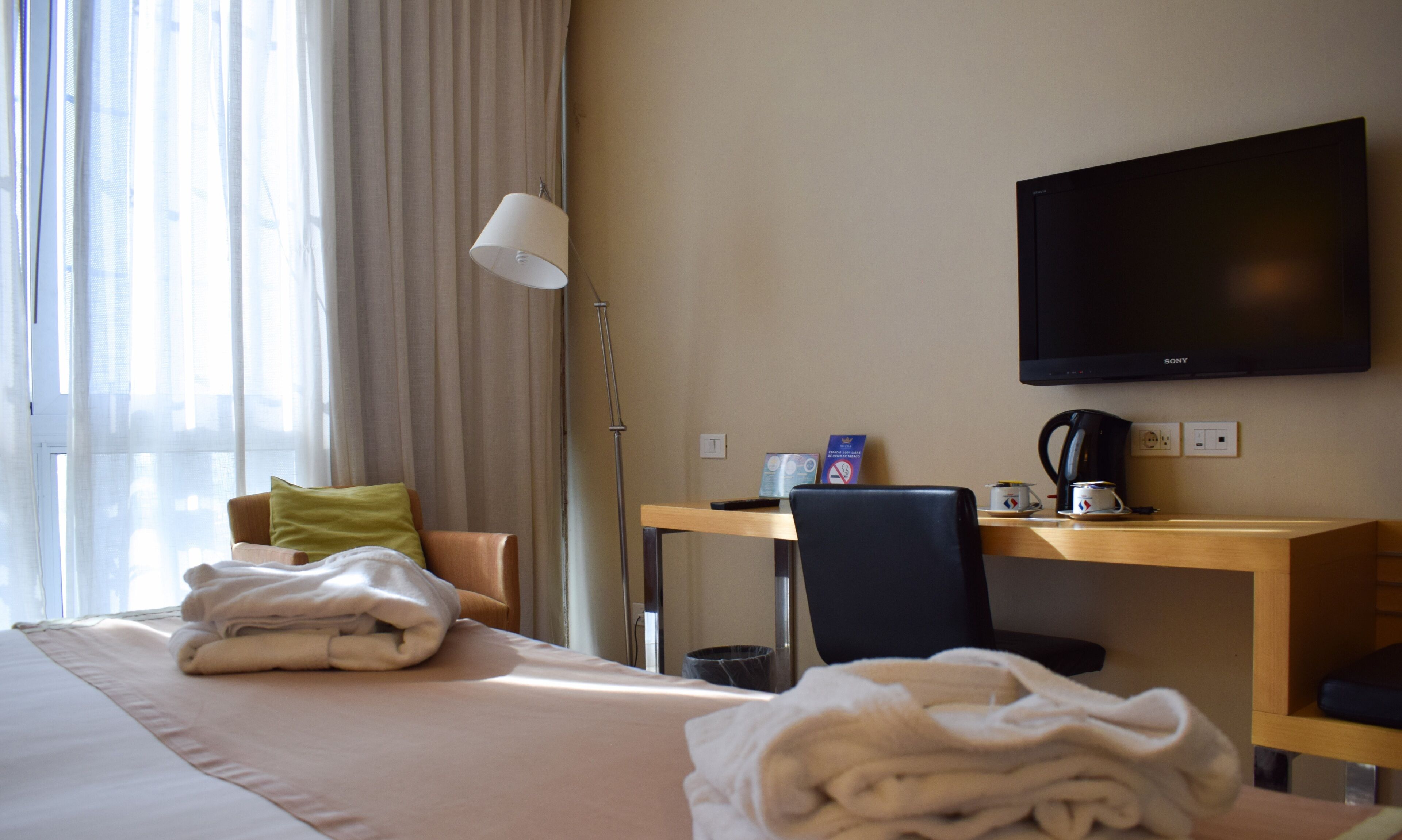 Habitación de hotel con cama, escritorio, silla, televisor y ventana con cortinas.