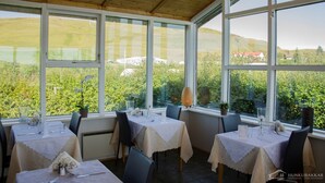 Un restaurant amb taules preparades per dinar, grans finestres que ofereixen vistes a zones verdes i edificis, i un sostre de fusta.