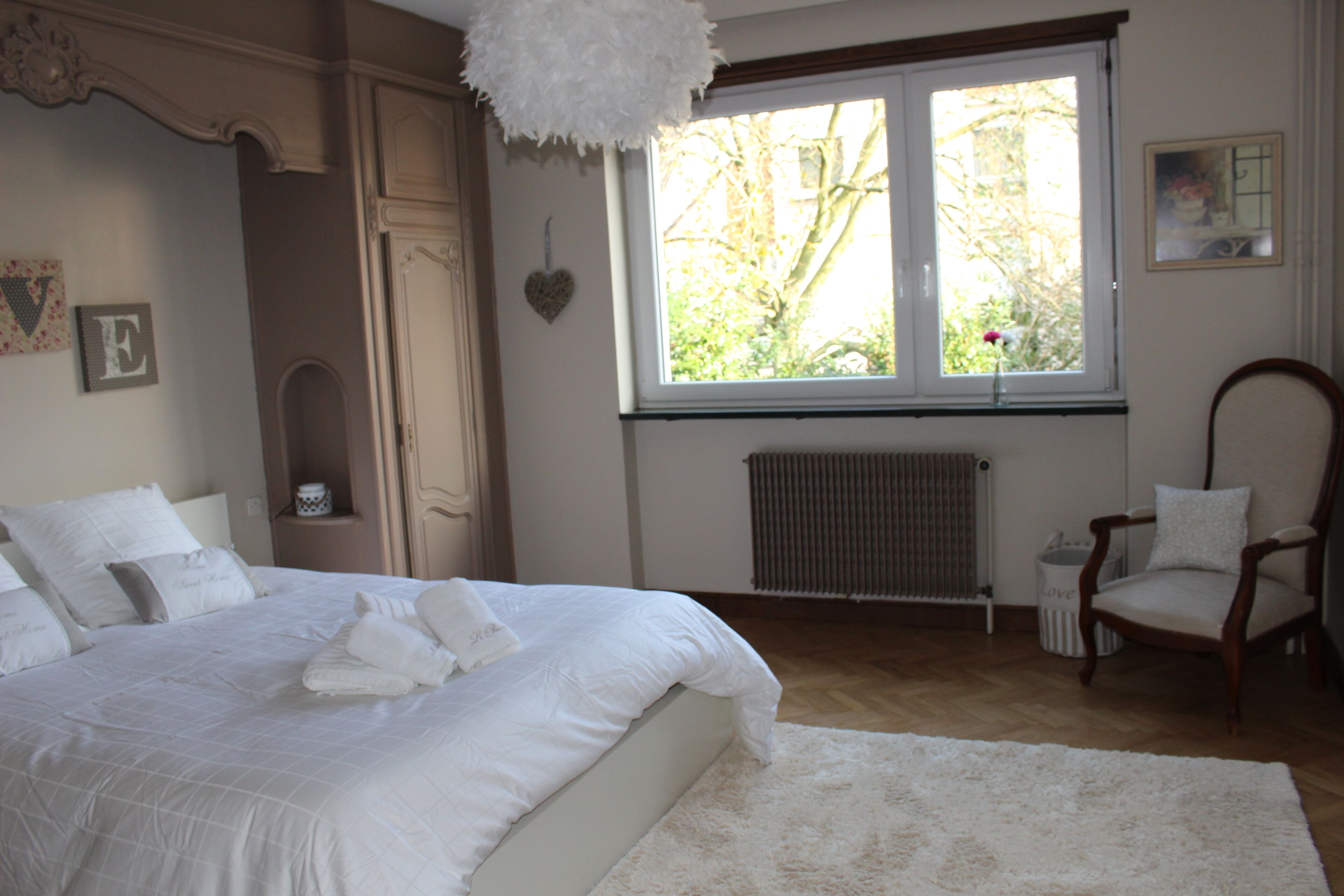 A bedroom with a large bed, a wooden chair, a radiator, and a window with a view of trees.