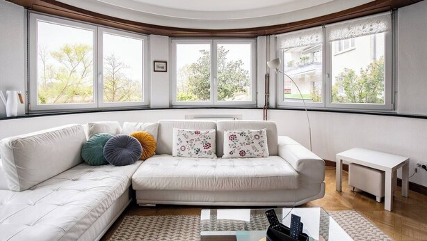 Una sala de estar moderna con un sofá blanco, cojines coloridos, una mesita blanca grande y ventanas que ofrecen vistas a árboles y edificios.